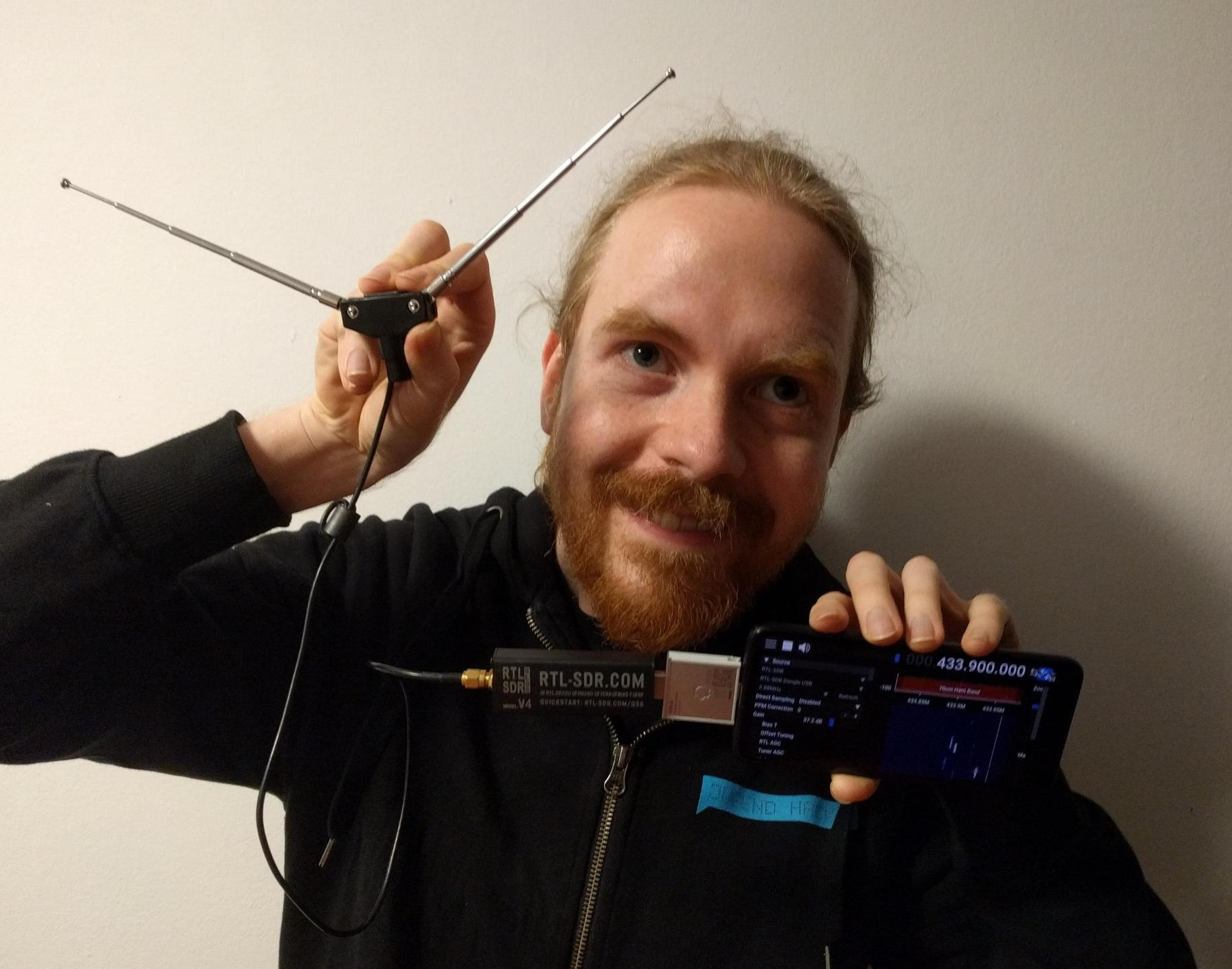 Me, a red-haired, red-bearded white human, hold a smartphone, connected to a Framework module, connected to my SDR stick, connected to an antenna! I look happy and tired.
